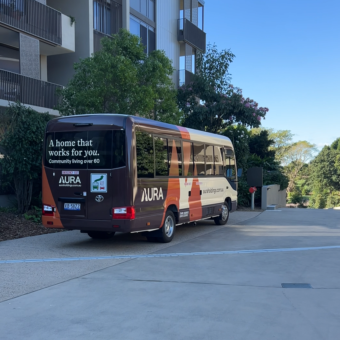 A brown and orange Aura minibus is parked outside a modern apartment building on a sunny day. The side of the bus displays text promoting community living for people over 60. Trees and greenery are in the background.