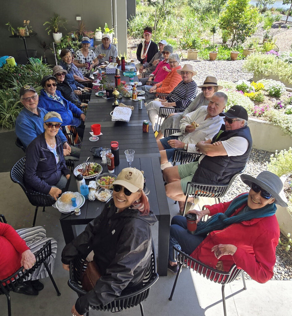 A group of people sits around a long outdoor table, enjoying food and drinks. They are smiling at the camera, wearing casual clothes, hats, and sunglasses, with plants and greenery in the background.